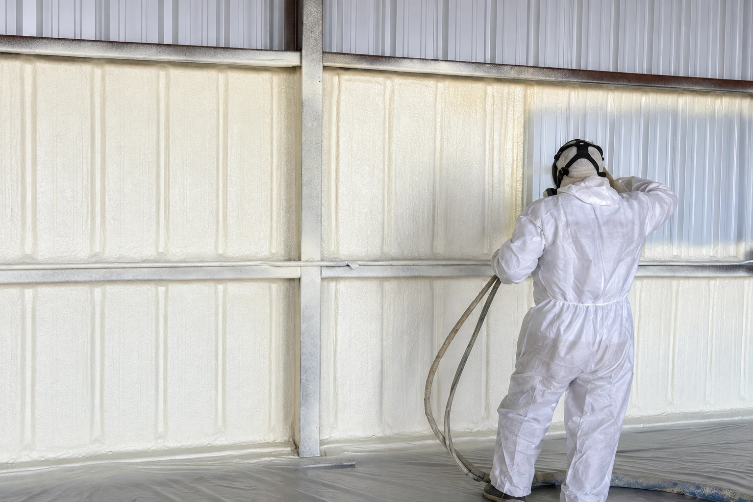 A worker in protective coveralls sprays white closed-cell foam insulation onto the interior wall of a metal building.
