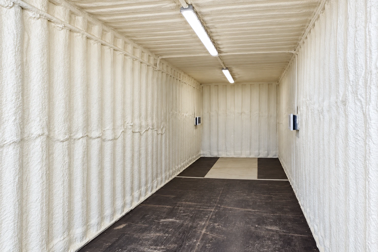 A long commercial container is lined with white spray foam insulation and lit by overhead lights.