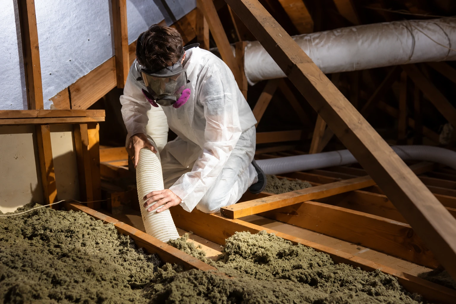 A worker in protective coveralls and a respirator uses a flexible hose to remove loose insulation from an attic.