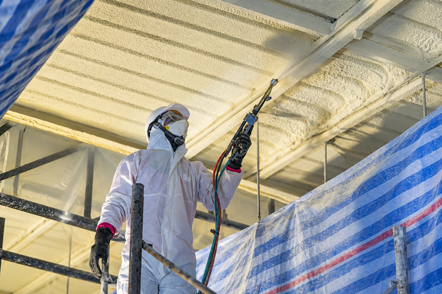A worker in protective coveralls and a respirator sprays foam insulation onto a commercial ceiling, with plastic sheeting around the work area.