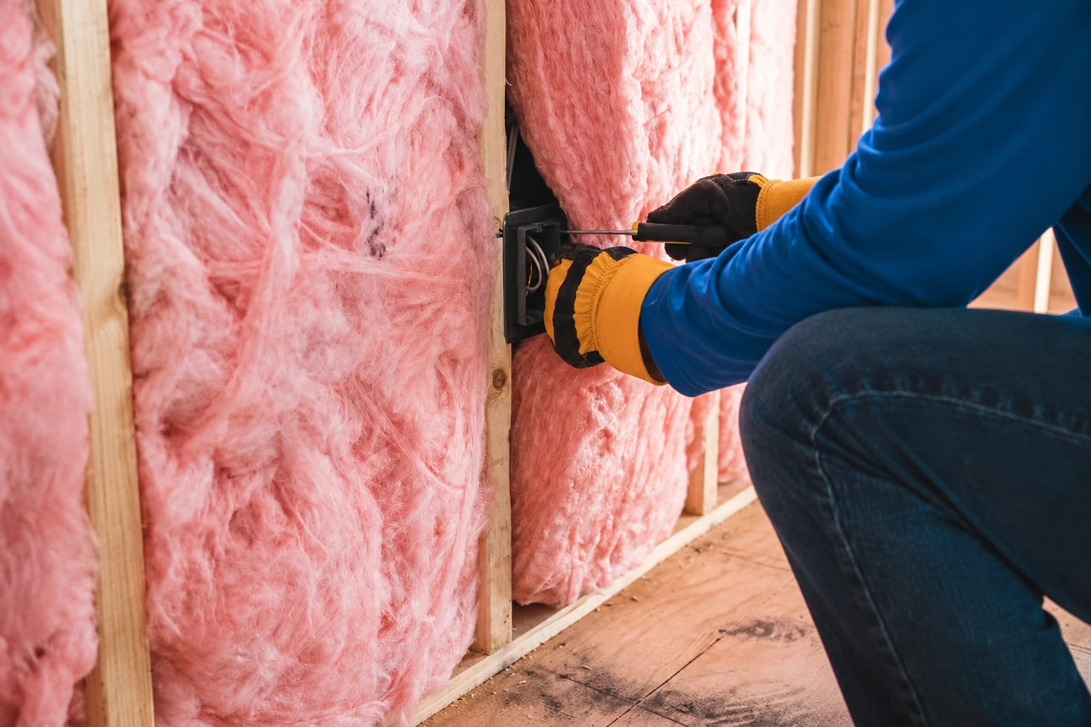 Worker in gloves adjusts an electrical box beside pink batt insulation fitted between wall studs.